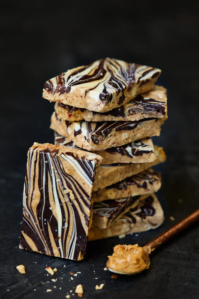 close up full size Double Chocolate Peanut Butter Bark stacked on a dark surface, featuring marbled chocolate swirls and chopped peanuts, with a spoonful of peanut butter beside i
