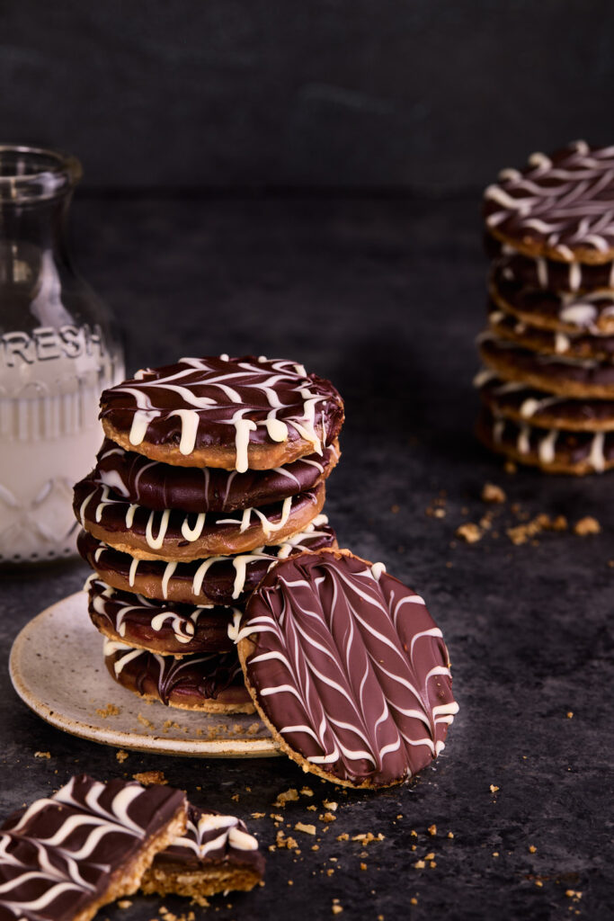 Stack of Chocolate Caramel Hobnobs with feathered dark and white chocolate topping, displayed on a plate with additional cookies in the background.