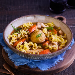 Bowl of homemade chicken soup with a clear golden broth, shredded chicken, carrots, egg noodles, and a matzoh ball, garnished with fresh dill and served on a wooden board with a blue cloth.