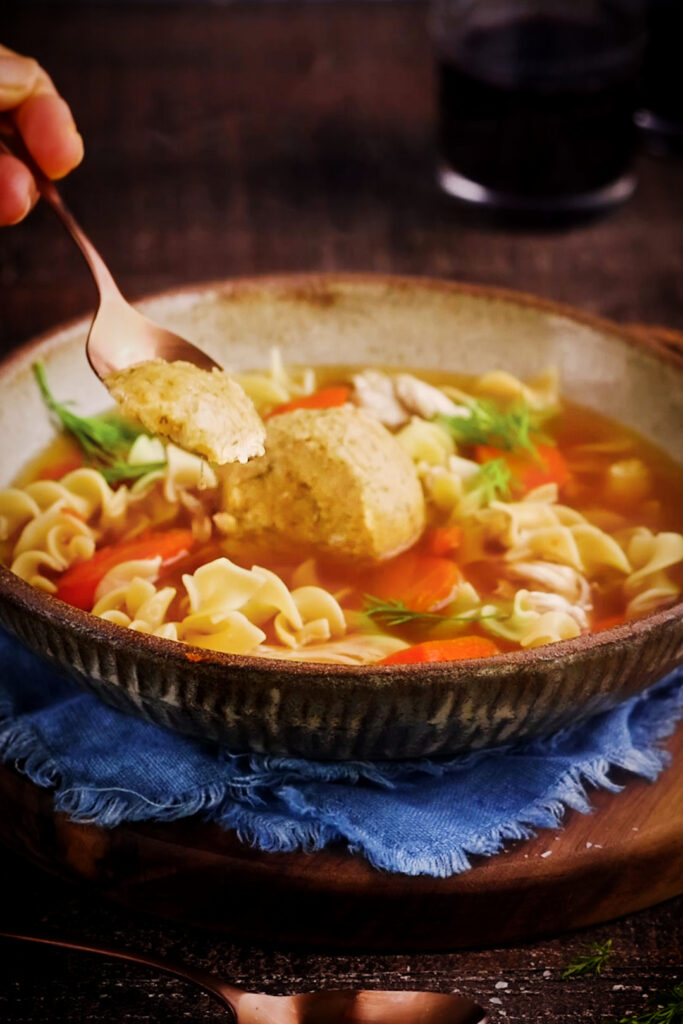 Close-up of a steaming bowl of homemade chicken noodle soup with egg noodles, shredded chicken, carrots, and fresh dill, featuring a fluffy matzoh ball in the centre as a spoon lifts a bite—perfect for “How to Make Fluffy Matzoh Balls (My Mom’s Secrets)”.