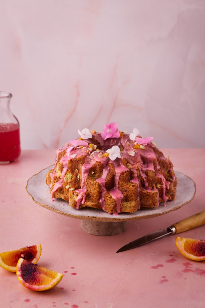 Blood Orange Coconut Bundt Cake with poppy seeds and pink blood orange glaze decorated with edible flowers on a cake stand.