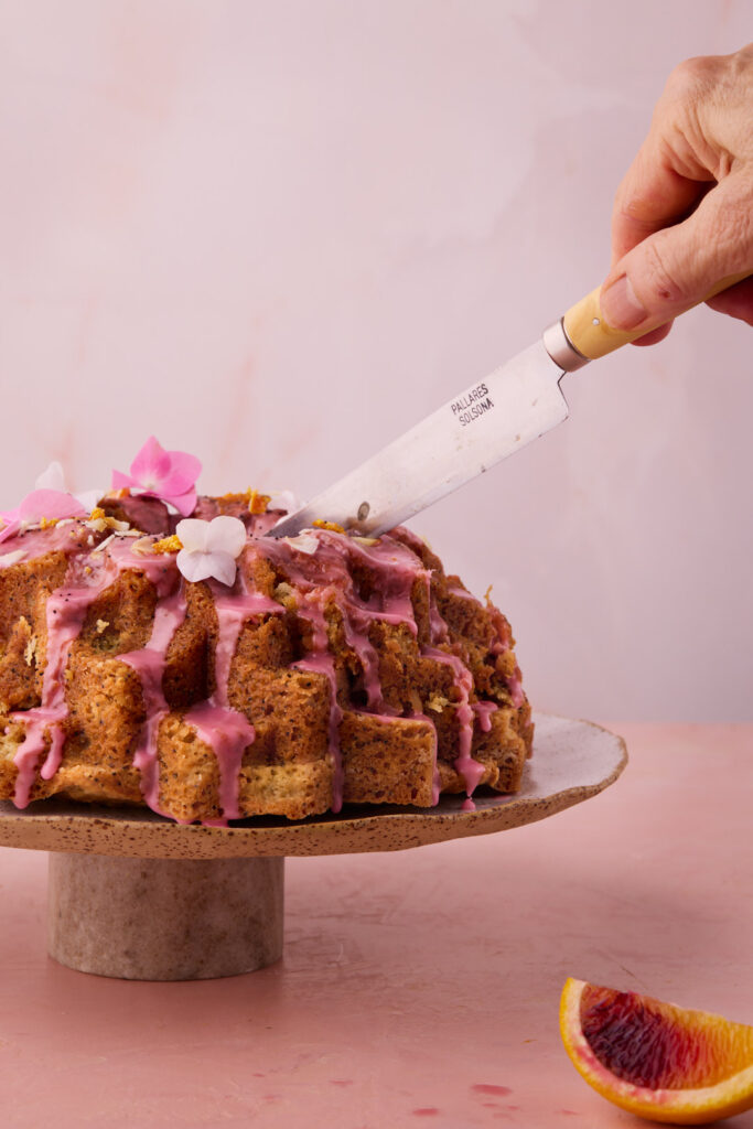 Knife slicing into Blood Orange Coconut Bundt Cake topped with pink blood orange glaze and edible flowers.