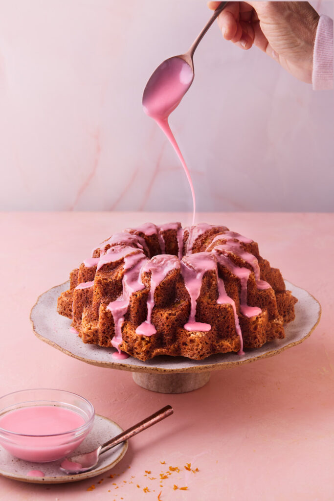 Pink blood orange glaze being poured over freshly baked Blood Orange Coconut Bundt Cake.