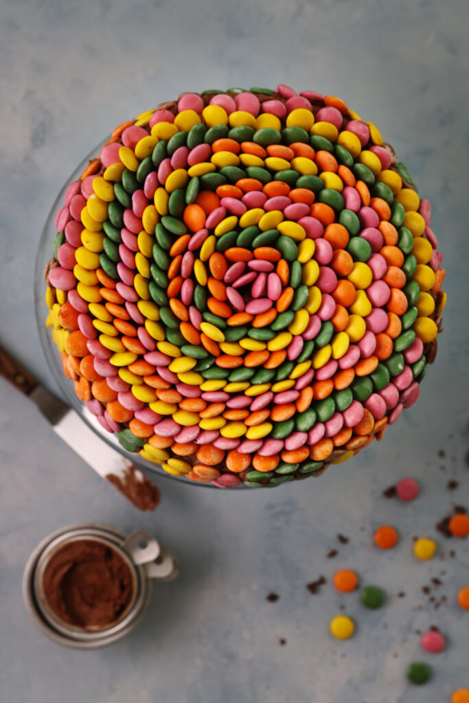 Overhead view of gluten free chocolate cake covered in Smarties arranged in a tight spiral pattern, showing yellow, orange, pink, and green candies on a blue surface.