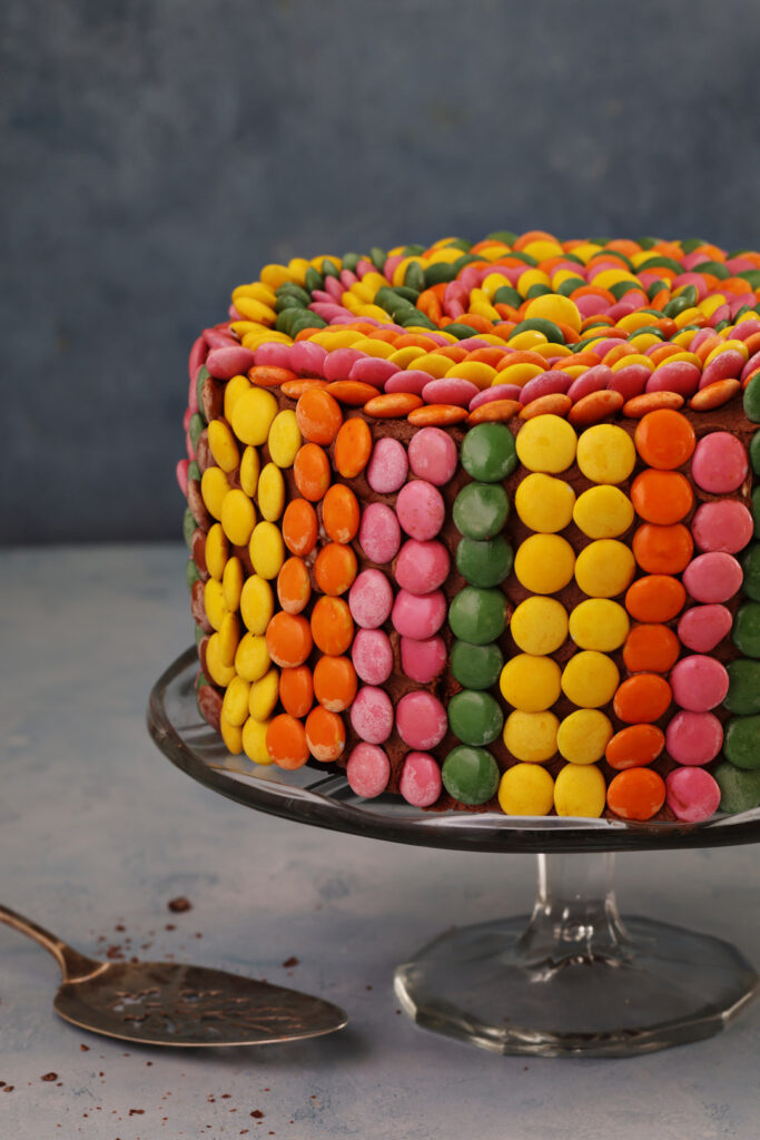 Side view of gluten free chocolate cake with mocha buttercream, decorated with vertical rows of colourful Smarties, showing texture and detail of the candy pattern.