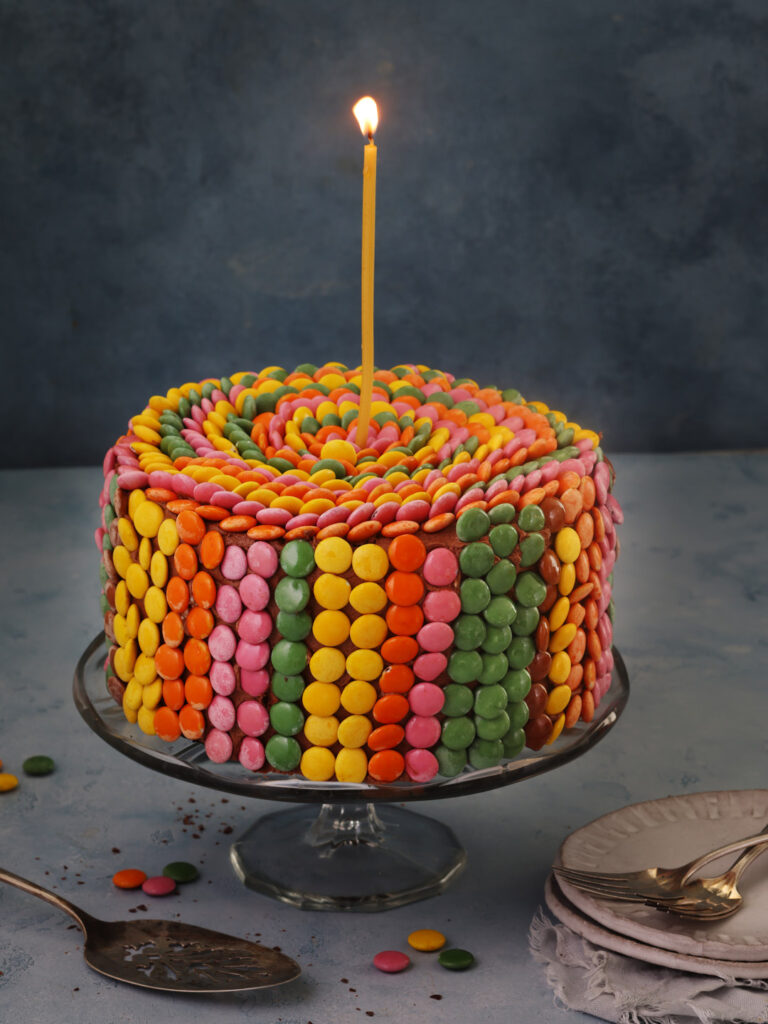 Gluten free chocolate cake with mocha buttercream decorated with colourful Smarties in a spiral pattern, topped with a lit birthday candle on a blue backdrop.