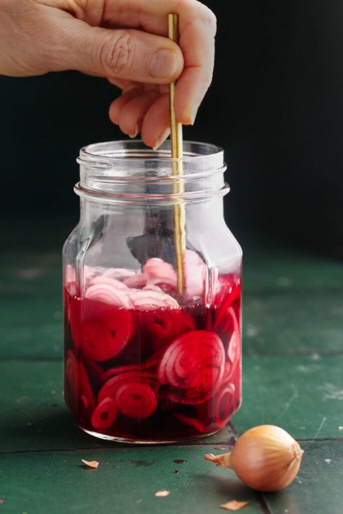 Jar of bright pink pickled shallots in brine being stirred with a gold spoon on a dark green surface.