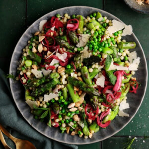 Overhead view of spring Israeli couscous salad with asparagus, peas, pickled shallots, toasted nuts, herbs, and shaved Parmesan on a dark plate