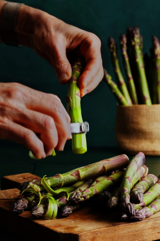 Hands peeling thick asparagus spears over a wooden cutting board, with fresh asparagus in the background for a spring salad preparation scene.