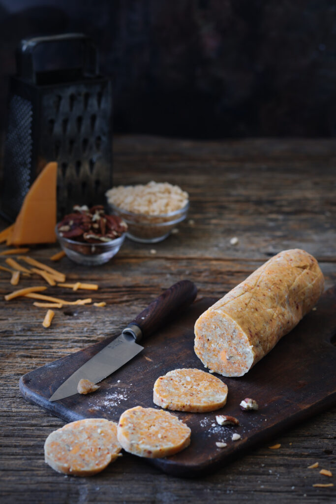 Log of savoury cheddar cheese biscuit dough sliced into rounds on a wooden cutting board, with cheddar cheese, pecans, and Rice Krispies in the background.