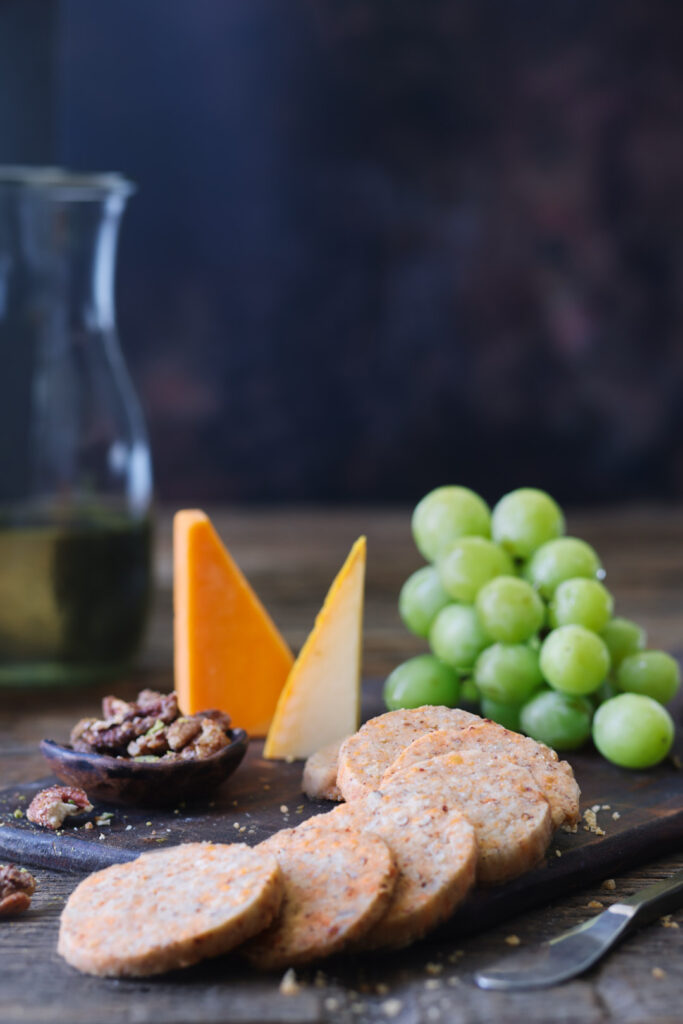Sliced savoury cheese biscuits served with aged cheddar, pecans, and green grapes on a rustic wooden board.