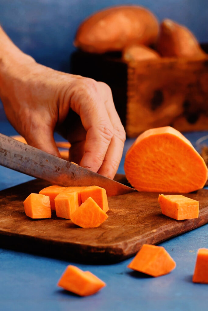 Hand cutting peeled sweet potato into cubes on a wooden cutting board for sweet potato breakfast waffles.